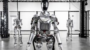 A photograph of a slim humanoid robot standing with reflective metal skin and a black motorcycle helmet